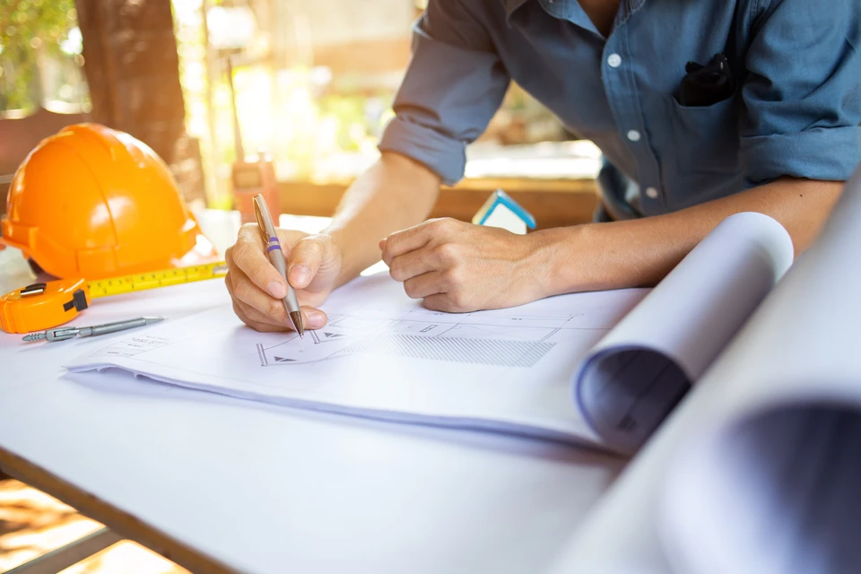 Professionnel en chemise bleue travaillant sur des plans architecturaux déroulés sur table, tenant un stylo pour annoter les documents